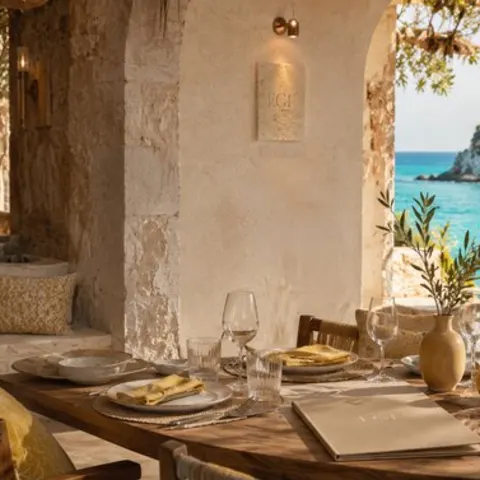 Stone arches over set wooden tables, an olive sprig in ceramic, the cove turquoise beyond