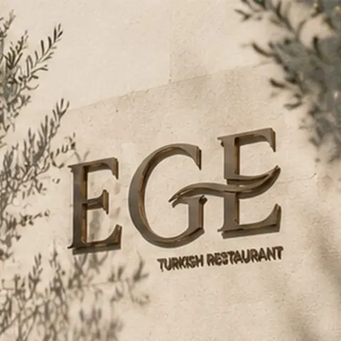 The EGE sign in brushed bronze on sunlit stone, olive shadows across the wall