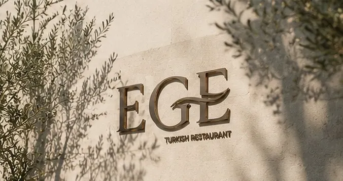 The EGE sign in brushed bronze on sunlit stone, olive shadows across the wall